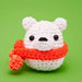 A white crocheted polar bear amigurumi toy wearing a bright orange-red knitted scarf, set against a solid green background.