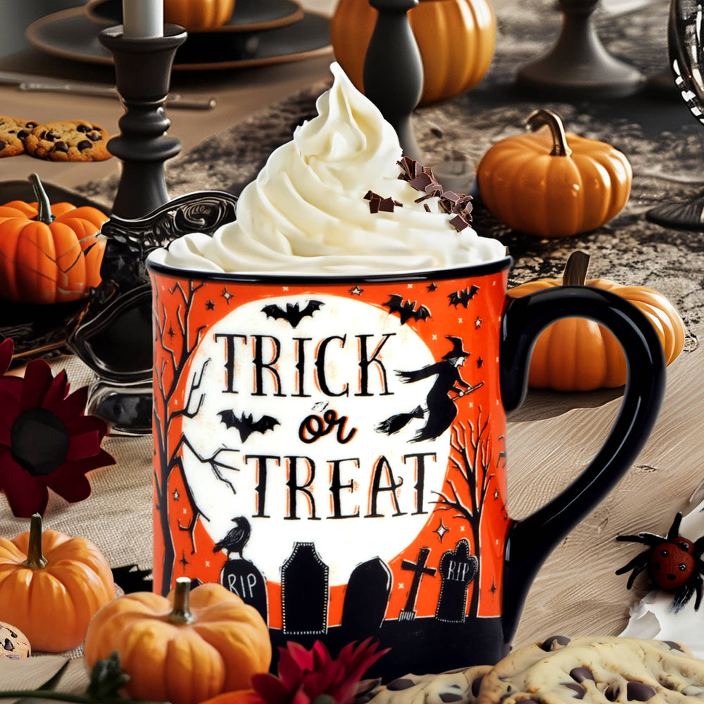 A Halloween-themed ceramic mug filled with whipped cream and chocolate shavings, set in a styled autumn table scene with pumpkins, candles, cookies, and a spider decoration. The mug features an orange and black design with a white moon and the words 'TRICK or TREAT' in black.