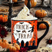 A Halloween-themed ceramic mug filled with whipped cream and chocolate shavings, set in a styled autumn table scene with pumpkins, candles, cookies, and a spider decoration. The mug features an orange and black design with a white moon and the words 'TRICK or TREAT' in black.