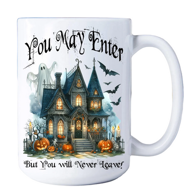 A white ceramic mug with a Halloween-themed graphic print featuring a haunted house, pumpkins, bats, a ghost, and the text 'You May Enter But You Will Never Leave!'.